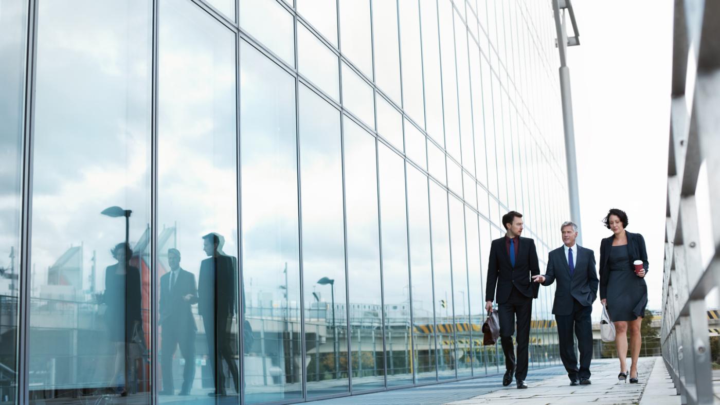 3 executive people walking together by the side of a build having glass walls.