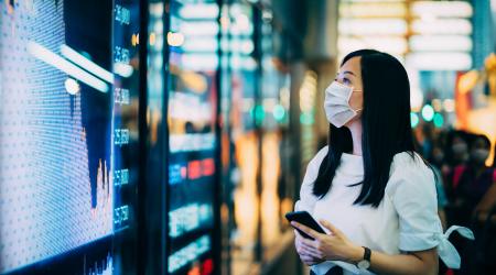 Woman in mask looks a television screen display stock tickers and graph